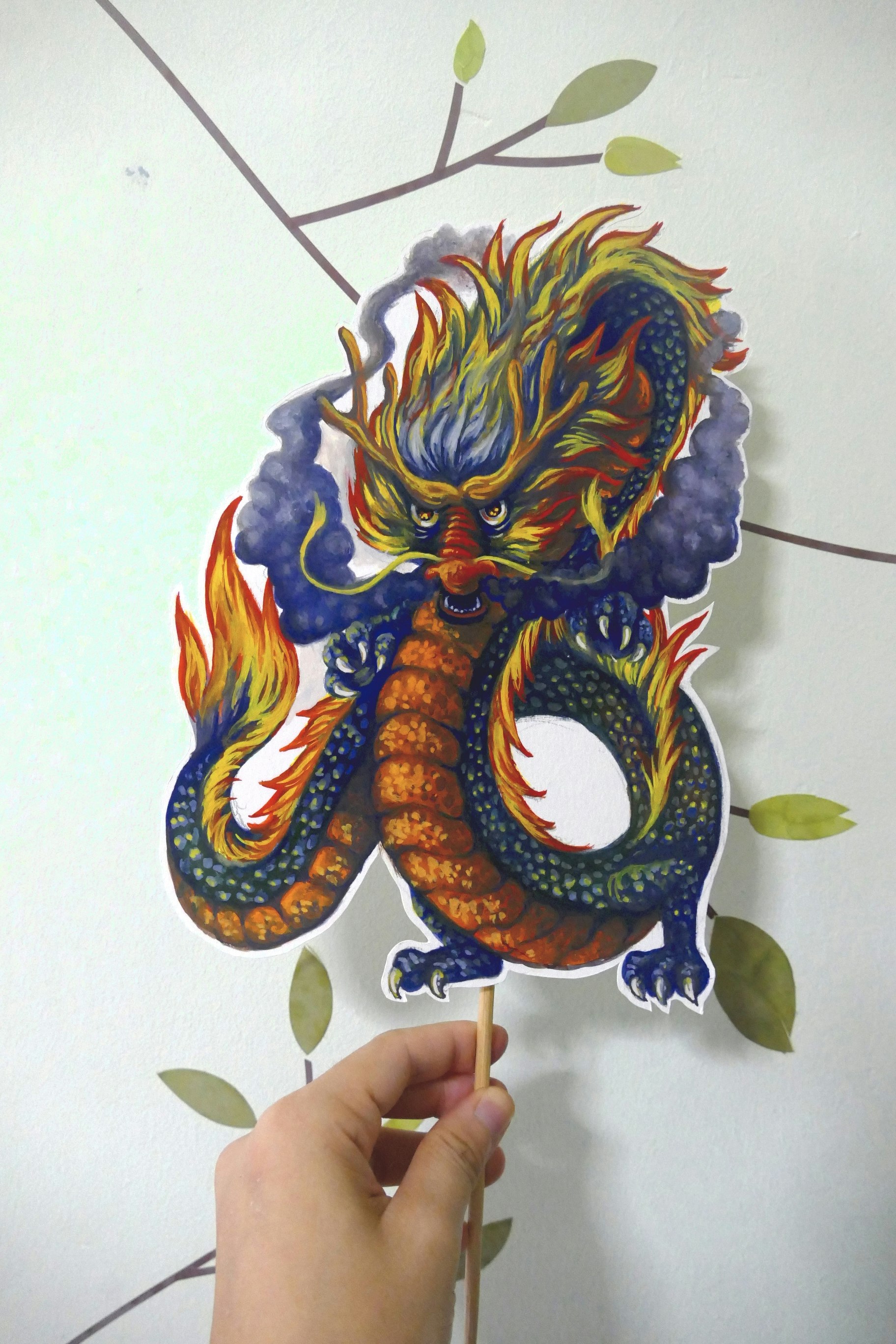 Poster painting cut-out on a stick of a menacing Chinese dragon with blue scales, a yellow belly, sharp claws, and a flaming yellow and red mane, with gray smoke bellowing out of its nostrils.