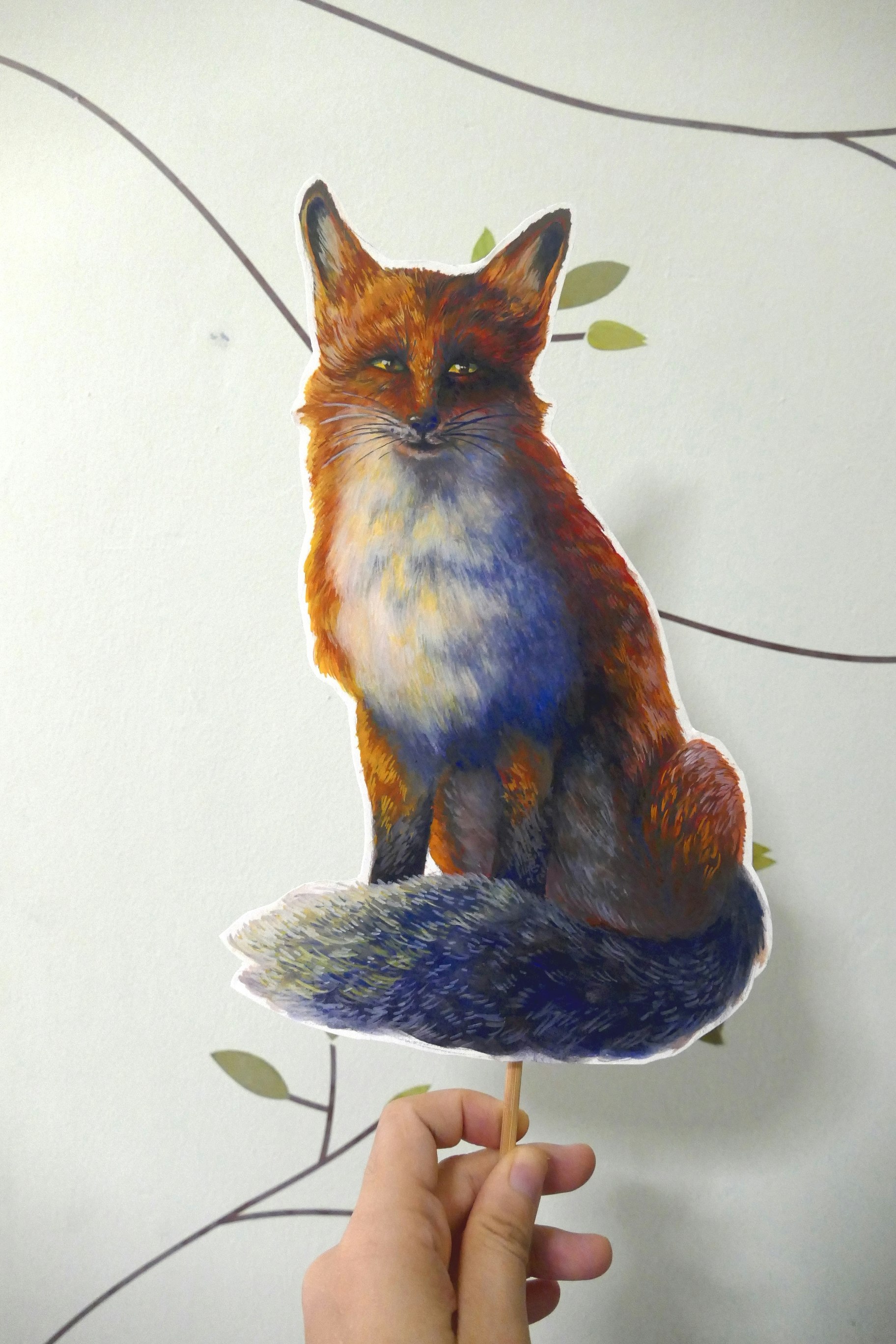Poster painting cut-out on a stick of a red fox sitting on its haunches. The shadows are a cool blue and its busy tail is curled around its legs.